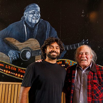 Thomas Scanlan, a young Indigenous man, with Kev Carmody, an older Indigenous man. They are standing in front of a large mural of Kev playing guitar.