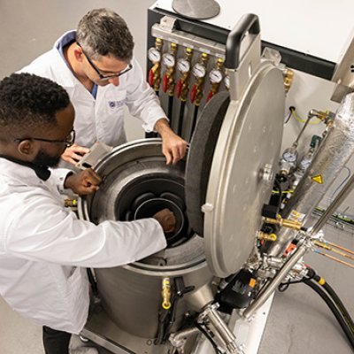 Two people wearing lab coats looking down into a round metal furnace